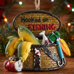 K&ouml;nnte beinhalten: Ein h&auml;ngendes Ornament in Form eines gelben Ziegelkorbs mit dem Text Hooked on FISHING. Es zeigt zwei gelbe Fische, eine rote Getr&auml;nkedose, eine rot-wei&szlig;e Angelpose, ein silbernes Werkzeug, einen gr&uuml;nen Beh&auml;lter und einen blauen Angelrutengriff.