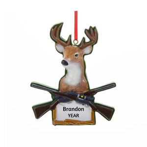 May include: A green and brown Christmas ornament featuring a deer head with crossed rifles and a personalized name tag that reads "Brandon YEAR".