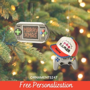 May include: Two Christmas ornaments featuring a video game theme. One ornament is a gray handheld game console with the words "Merry Xmas" on the screen. The other ornament is a gray video game controller with a red and white baseball cap on top that says "Pro Gamer".