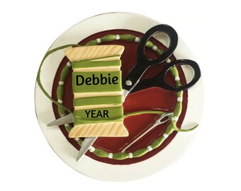 May include: A white ceramic plate with a red and green border. The plate features a spool of thread with the name "Debbie" and the word "YEAR" on it. A pair of black scissors and a needle are also on the plate.