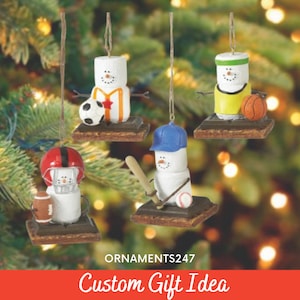 以下が含まれることがあります： マシュマロの人形の形をしたクリスマスオーナメントが4つ。それぞれ異なるスポーツのユニフォームを着て、スポーツボールやバットを持っています。オーナメントはクリスマスツリーに飾られています。