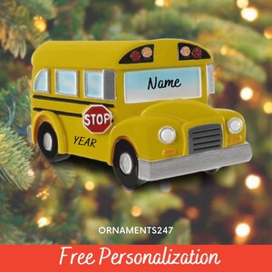 Puede incluir: Adorno de autobús escolar amarillo con detalles en negro, un parachoques gris y una señal de stop roja. El autobús tiene la palabra "Name" en la ventana delantera y "STOP" y "YEAR" en el lateral. El adorno está sobre un fondo borroso.
