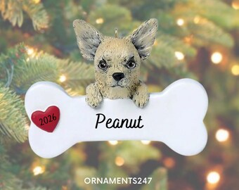 May include: A beige and black speckled dog ornament featuring a sculpted head and paws resting on a white bone-shaped plaque. The plaque displays the name Peanut in black script and a small red heart with the year 2026 printed in black ink.