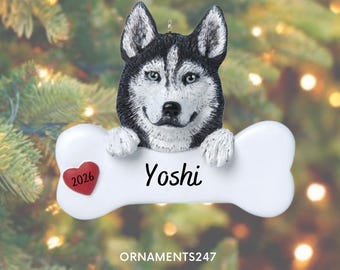 May include: A hanging Christmas ornament featuring a black and white Siberian Husky dog head resting on a white bone-shaped plaque. The plaque displays the name Yoshi in black script and a small red heart with the year 2026 printed on it.
