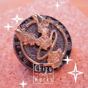 Puede incluir: Un pin de esmalte dorado y negro con un fénix estilizado con las alas extendidas, una rosa y un diseño circular con intrincados detalles. El pin dice "Chizu Works" en la parte inferior.