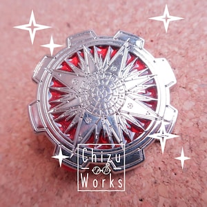 May include: A silver and red enamel pin with a starburst design and intricate details. The pin features a circular shape with a gear-like border and a central starburst with a red enamel background. The text "Chizu Works" is visible at the bottom of the pin.