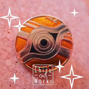 May include: A gold-colored metal pin with an orange eye design. The eye is surrounded by a black ring and a wavy line. The pin is circular and has a shiny finish. The text "Chizu Works" is printed below the eye.