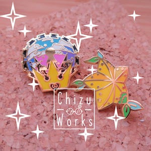 May include: Two enamel pins, one with a gold crown and a blue, white and pink sky with stars, and the other with a yellow star with green leaves. The text "Chizu Works" is visible below the pins.