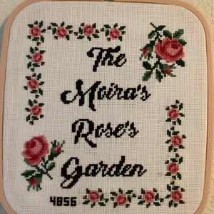May include: A white cross-stitch embroidery with a pink and green floral border. The text "The Moira's Rose's Garden" is stitched in black thread. The number "4856" is stitched in the bottom left corner.
