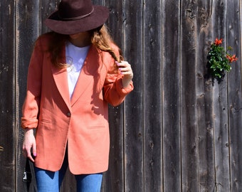 May include: A woman wearing a coral blazer with a brown fedora hat. She is standing in front of a wooden fence.