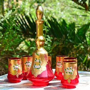 May include: A set of six red glass tumblers with gold accents and white floral designs, and a matching red glass decanter with a gold stopper.