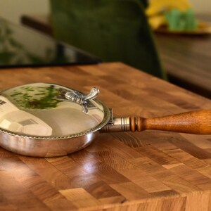 May include: A silver serving dish with a wooden handle. The dish has a domed lid with a decorative knob. The dish is sitting on a wooden surface.