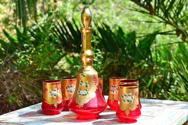 May include: A set of six red glass tumblers with gold accents and white floral designs, and a matching red glass decanter with a gold stopper.