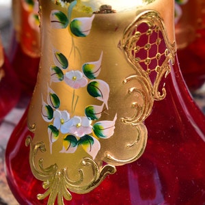 May include: Set of four red glass decanters with gold painted floral designs and accents. The decanters have a pear-shaped body and a round base.