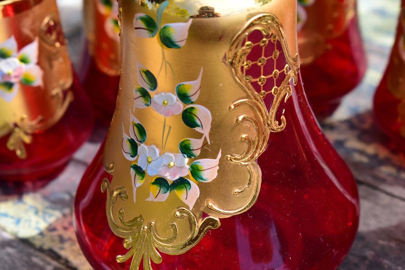 May include: Set of four red glass decanters with gold painted floral designs and accents. The decanters have a pear-shaped body and a round base.