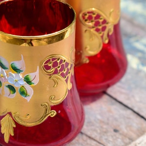 May include: Set of three red glass tumblers with gold trim and hand-painted floral designs. The glasses have a flared base and a wide mouth.