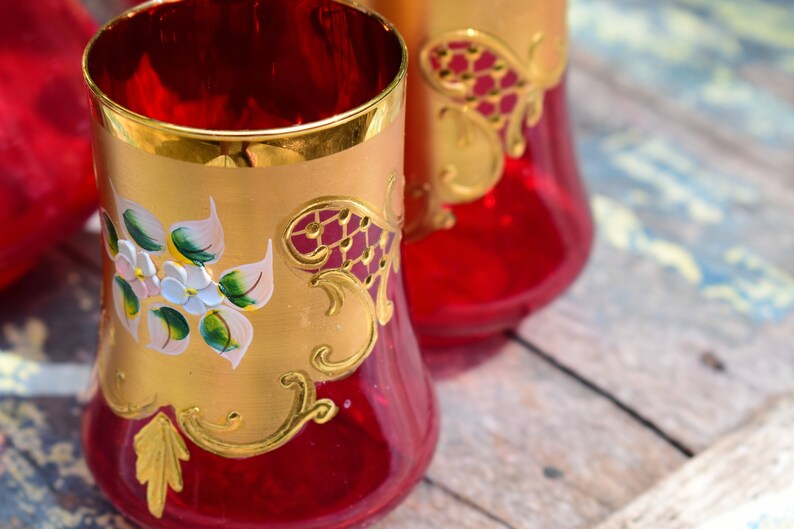 May include: Set of three red glass tumblers with gold trim and hand-painted floral designs. The glasses have a flared base and a wide mouth.