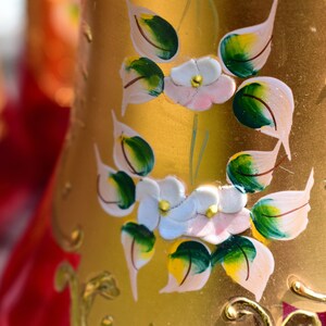 May include: A gold-colored glass bottle with a red base and painted floral designs. The flowers are white with pink centers and green leaves. The bottle has a gold-colored trim.