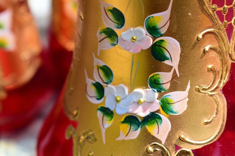 May include: A gold-colored glass bottle with a red base and painted floral designs. The flowers are white with pink centers and green leaves. The bottle has a gold-colored trim.