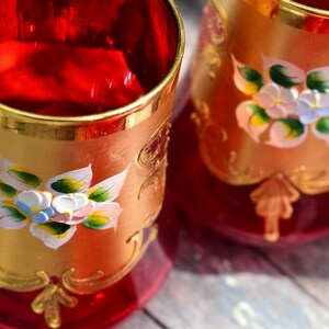 May include: Two red glass cups with gold rims and painted floral designs. The cups have a gold and red pattern on the bottom.