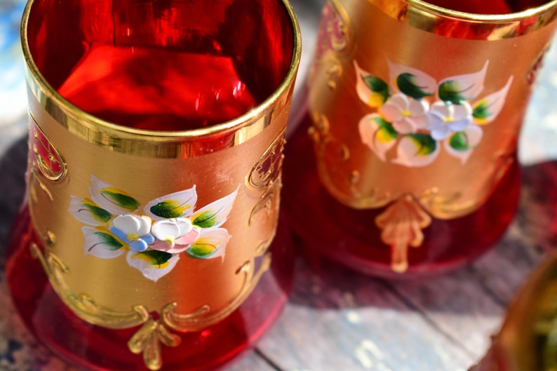 May include: Two red glass cups with gold rims and painted floral designs. The cups have a gold and red pattern on the bottom.
