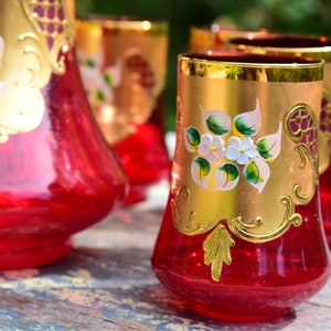 May include: Set of four red glass tumblers with gold accents and floral designs. The glasses are decorated with white flowers and green leaves.
