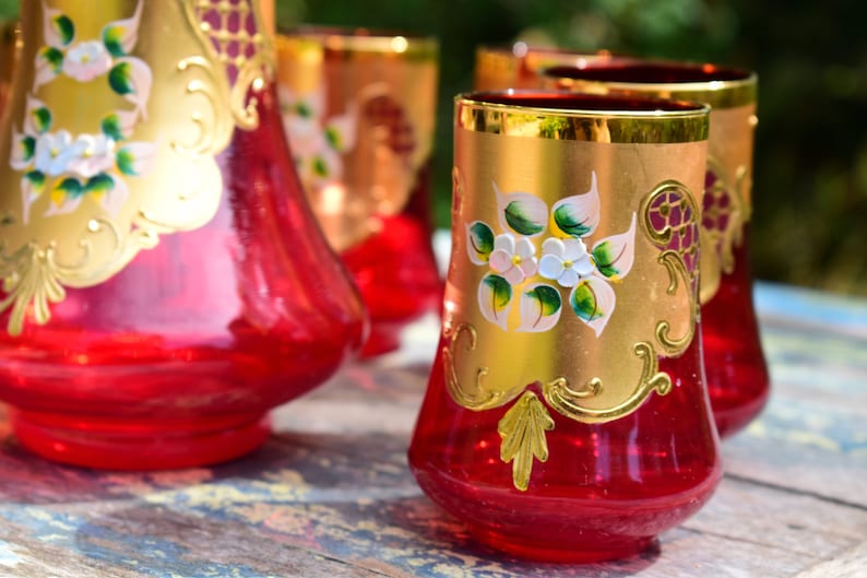 May include: Set of four red glass tumblers with gold accents and floral designs. The glasses are decorated with white flowers and green leaves.