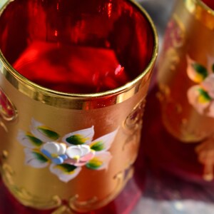 May include: Two red glass cups with gold rims and painted floral designs. The cups are decorated with white, yellow, and green flowers.