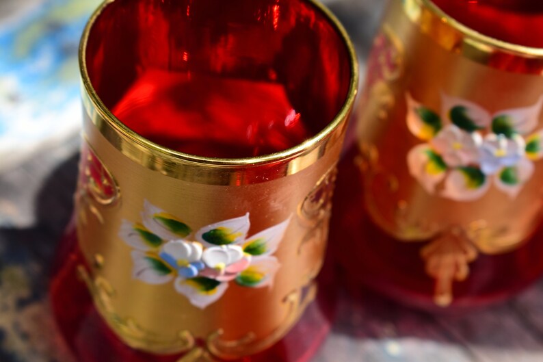 May include: Two red glass cups with gold rims and painted floral designs. The cups are decorated with white, yellow, and green flowers.