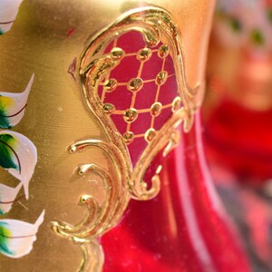 May include: A red glass vase with gold painted floral designs and a gold-painted geometric pattern. The vase has a bulbous shape and a narrow neck.