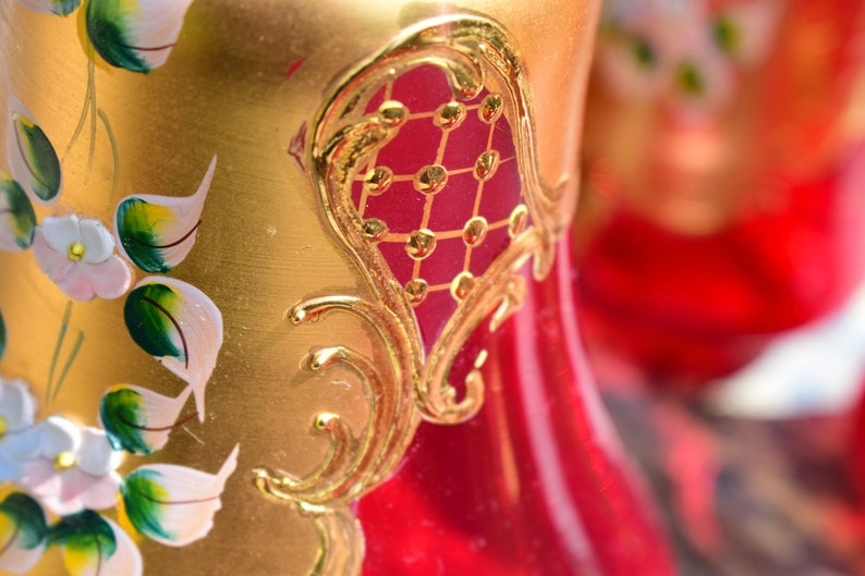 May include: A red glass vase with gold painted floral designs and a gold-painted geometric pattern. The vase has a bulbous shape and a narrow neck.