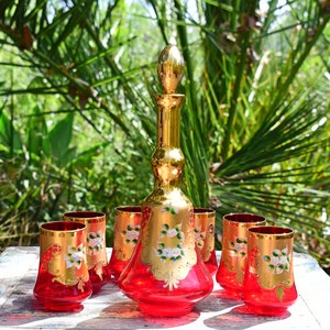 May include: A set of six red glass tumblers and a matching decanter with gold accents and floral designs. The glasses and decanter are on a white wooden table.