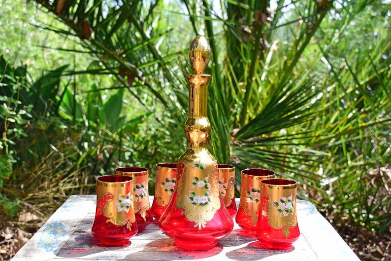 May include: A set of six red glass tumblers and a matching decanter with gold accents and floral designs. The glasses and decanter are on a white wooden table.