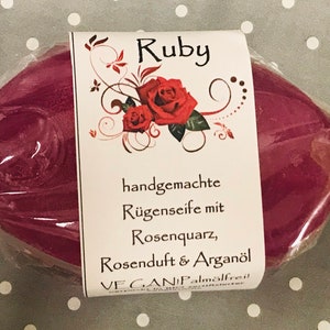 Könnte beinhalten: Ein rubinrotes, ovales Seifenstück in klarer Verpackung. Das Etikett trägt die Aufschrift "Ruby" mit floralem Design und deutschem Text. Die Seife ist handgemacht, vegan und enthält Rosenquarz, Rosenduft und Arganöl.