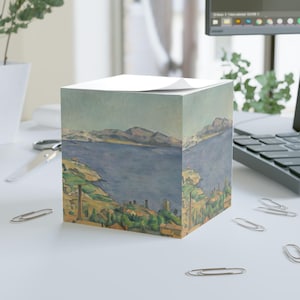 May include: A cube-shaped notepad featuring a landscape painting with a blue lake, green hills, and distant mountains. The artwork is reminiscent of a classic painting style, with soft colors and visible brushstrokes. The notepad sits on a white surface.