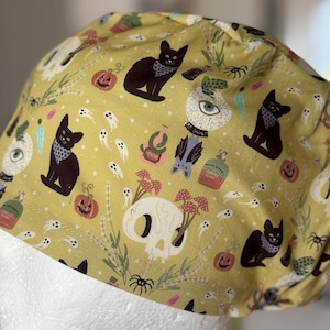 Può includere: Una cuffia chirurgica in tessuto giallo senape con un motivo ripetuto di gatti neri con bandana, teschi bianchi con funghi, piccoli fantasmi, zucche arancioni, sfere di cristallo, pipistrelli, ragni e varie boccette di pozioni magiche.
