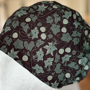 Può includere: Un cappello marrone scuro con un motivo ripetuto di foglie verdi e zucche verde chiaro. Il cappello è fatto di tessuto ed è progettato per adattarsi alla testa. Il design è una stampa botanica.