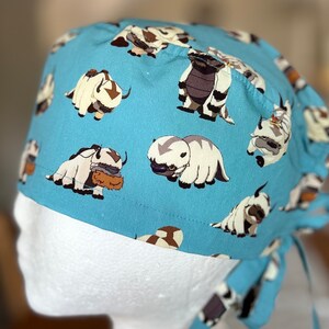 May include: A blue scrub hat with a white tie, featuring a repeating pattern of cartoon white and brown yak-like creatures.