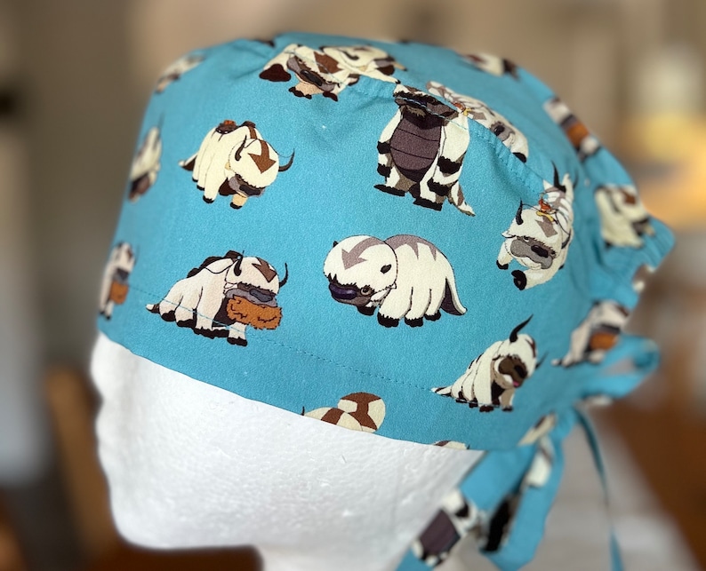 May include: A blue scrub hat with a white tie, featuring a repeating pattern of cartoon white and brown yak-like creatures.