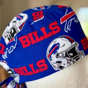 Może przedstawiać: Niebieski czepiec chirurgiczny z nadrukiem Bills. Czapka przedstawia logo drużyny, kaski do futbolu amerykańskiego i słowo "BILLS" w kolorze czerwonym. Czapka jest wykonana z miękkiej, prawdopodobnie bawełnianej tkaniny.
