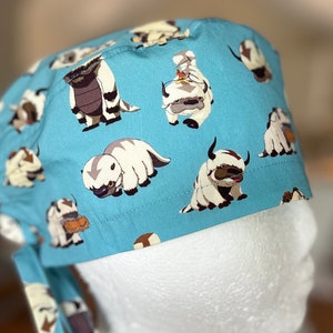 May include: A blue scrub hat with a white and brown patterned design featuring cartoon images of a furry creature.