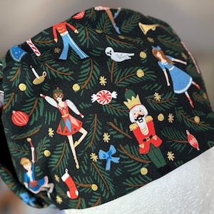 Op de afbeelding: Zwarte scrubcap met een feestelijk Notenkraker-thema. De cap toont illustraties van ballerina's, tinnen soldaten, zuurstokken en kerstboomtakken. Gouden sterren en accenten geven het ontwerp een vleugje glans.