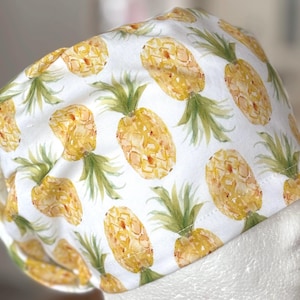 Puede incluir: Gorro quirúrgico blanco con un patrón repetido de piñas en acuarela amarilla y verde. El gorro está diseñado para cubrir la cabeza, comúnmente utilizado en entornos médicos.