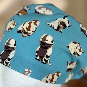 Peut inclure: Un bonnet de chirurgien bleu avec un motif r&eacute;p&eacute;t&eacute; de cr&eacute;atures blanches et poilues avec des marques brunes. Les cr&eacute;atures sont stylis&eacute;es pour ressembler &agrave; un animal mythique.