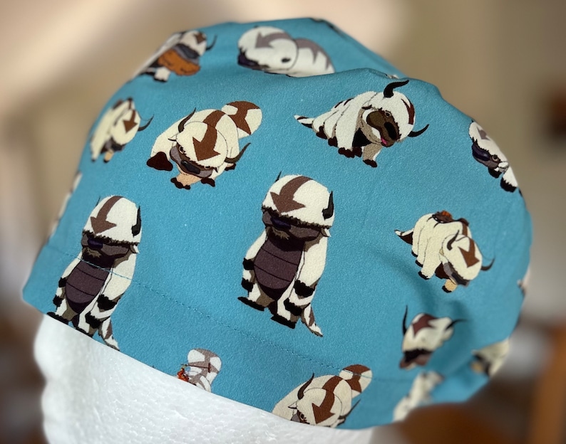 Peut inclure: Un bonnet de chirurgien bleu avec un motif r&eacute;p&eacute;t&eacute; de cr&eacute;atures blanches et poilues avec des marques brunes. Les cr&eacute;atures sont stylis&eacute;es pour ressembler &agrave; un animal mythique.