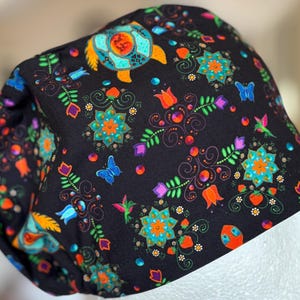 May include: A black surgical scrub cap with a colorful floral and butterfly pattern. The design includes orange, teal, and purple flowers, butterflies, and a turtle. The cap is made of a soft, patterned fabric.