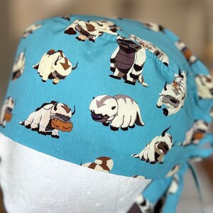 May include: A blue surgical scrub hat with a white and brown patterned design of cartoon yak-like creatures. The hat has a tie at the back.