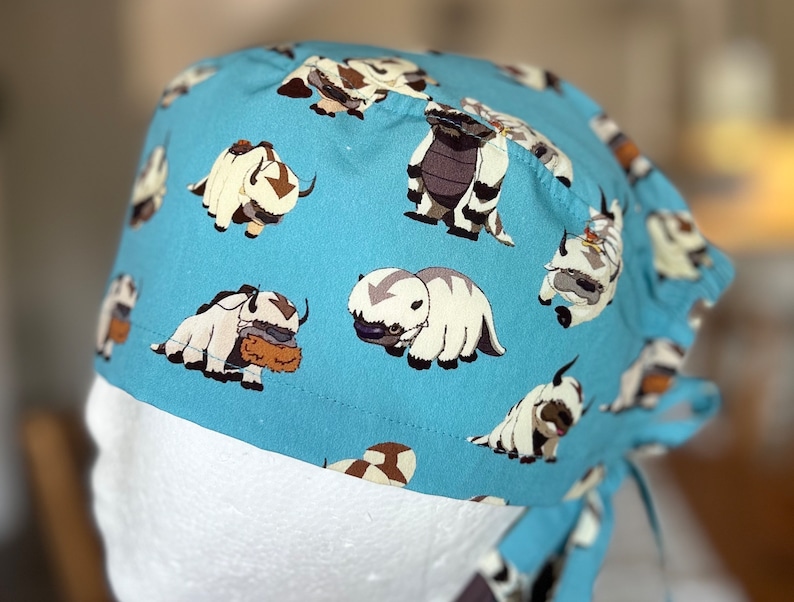 May include: A blue surgical scrub hat with a white and brown patterned design of cartoon yak-like creatures. The hat has a tie at the back.