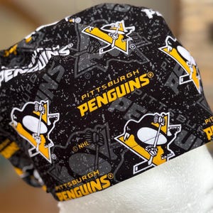May include: A black and gray patterned cap featuring the Pittsburgh Penguins logo in yellow and white. The cap has the words "PITTSBURGH PENGUINS" printed on it. The cap is made of a soft, flexible material.