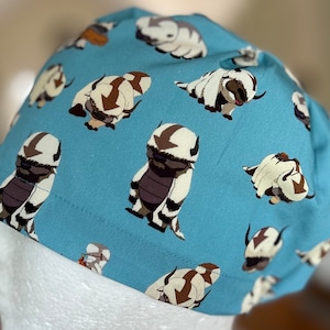 Peut inclure: Un chapeau en tissu bleu avec un motif r&eacute;p&eacute;t&eacute; d'animaux de dessin anim&eacute; blancs et bruns. Les animaux sont stylis&eacute;s et ressemblent &agrave; une cr&eacute;ature mythique avec une longue crini&egrave;re flottante.